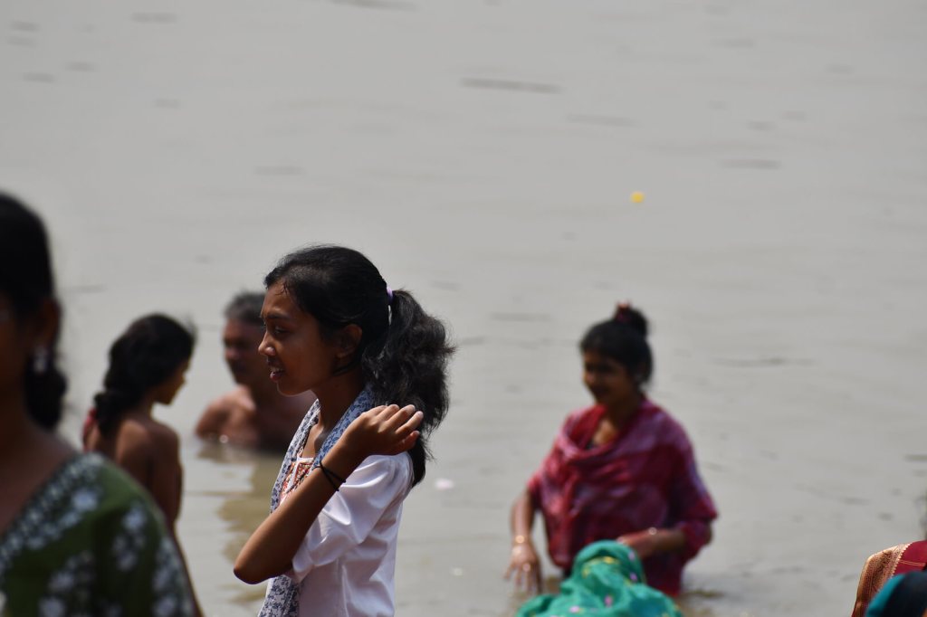 devotees bathing at the Dakshineswar Kali Temple in Kolkata, with the Ganga River and the Howrah Bridge in the background. The temple is a major pilgrimage site in West Bengal, attracting millions of visitors each year for its open holy bath.
